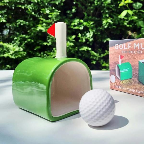 A green golf-inspired ceramic mug placed on its side to act as a putting hole next to a white golf ball.