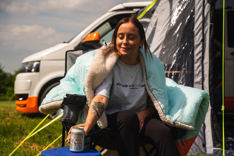 A young lady sat in a camping chair enjoying a drink whilst camping wrapped in an Insulated Outdoor Blanko by OLPRO in Tonal Treetops design. imageset:Tonal Tree Tops