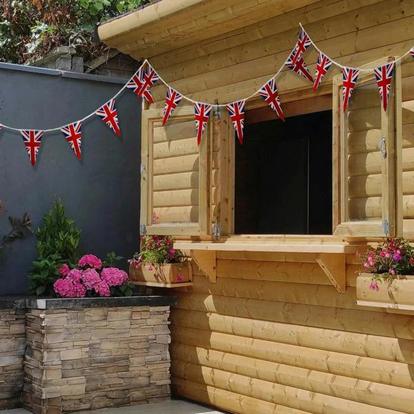 Union Jack flag bunting in vibrant red, white, and blue, strung across a wooden cabin window, enhancing a garden setting