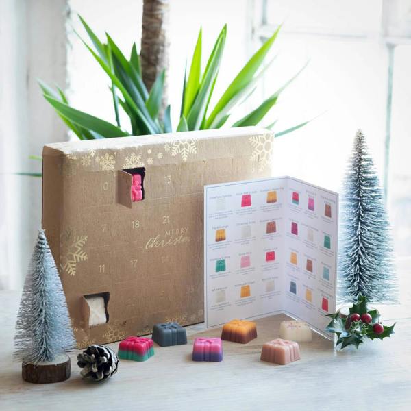 A kraft-paper advent calendar with gold snowflake details and an integrated stand, showing open windows with little gift-box-shaped wax melts, displayed with a scent recipe card, miniature pine trees, and a pinecone.