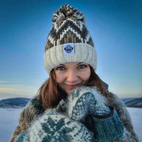  Set against a beautiful blue sky in Norway woman is wearing a Super Bobble hat in cream and soft brown shades in a zig-zag design, The hat  has a deep ribbed headband, contains reflective yarns and has a luxurious sherpa fleece lining.