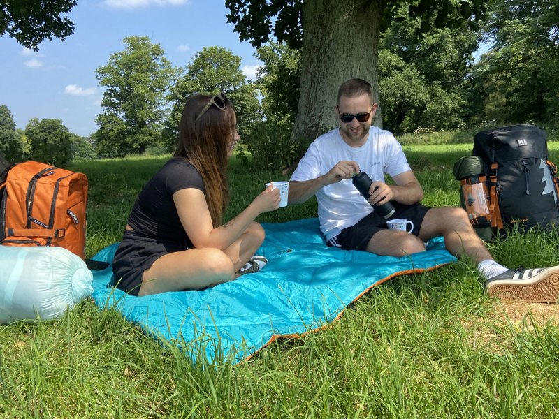 Two adults using the Original Insulated Outdoor Reversible Blanko from OLPRO as  a picnic blanket sat in a field with a treeline in the background.