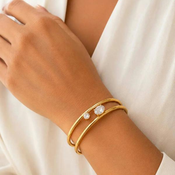 A close-up of a person's wrist wearing two thin gold bangles, one with a small clear stone, against a soft white garment background.