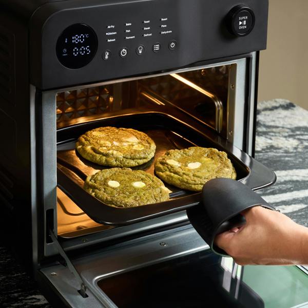 A person wearing a black oven mitt pulls out a tray with three large green cookies from a Fridja f66 Super Oven - 15L Digital Air Fryer Oven, whose display shows 18:05.