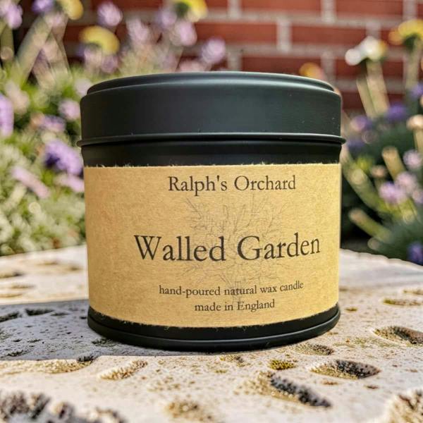 A black metal candle tin with a kraft paper label reading 'Ralph's Orchard' and 'Walled Garden', photographed outdoors on a stone surface with blurred flowers and a brick wall in the background.