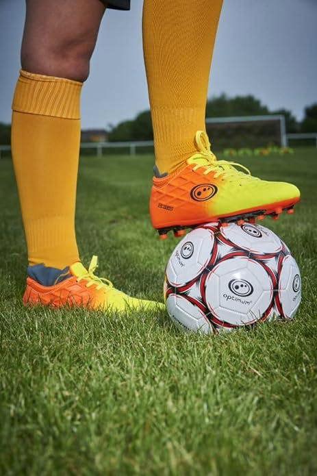 Player in orange Optimum Ignisio boots and orange socks resting foot on a football on a grass pitch - Optimum