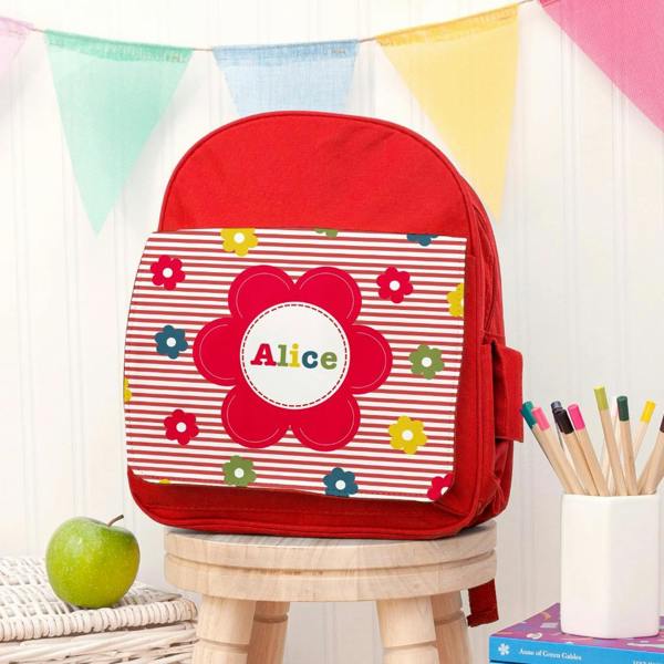 Personalised bright red girl's floral backpack custom printed with the name Alice on a wooden stool next to coloured pencils.