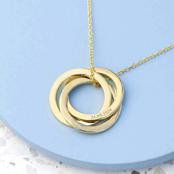 A gold personalised Russian ring necklace with three interlocking bands, one engraved with the date 24.09.2024, resting on a pastel blue circular surface.