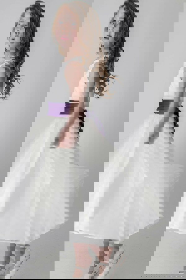 Young girl wearing an ivory dress with a purple belt on a plain background
