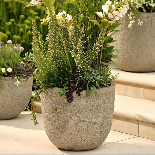 Cranford round garden planters, shown in a rustic, textured ficonstone finish resembling weathered light grey stone. The planters feature varied summer plantings including alliums and ornamental grasses, set upon stone patio steps in a sun-drenched outdoor living area. The aesthetic leans heavily into modern coastal garden styling and al fresco dining decor.