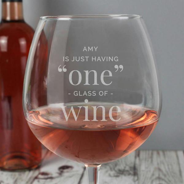 An extra-large clear glass filled with rosé wine up to the halfway mark, featuring white etched lettering that reads AMY IS JUST HAVING "one" GLASS OF wine, with a full wine bottle blurred in the background.