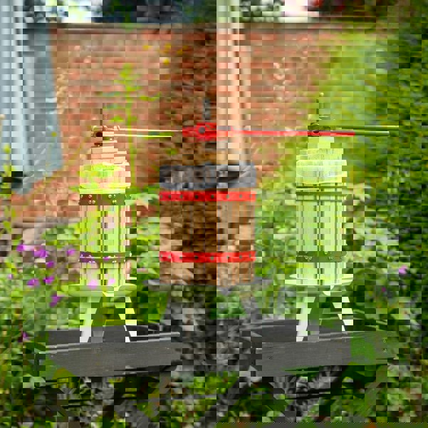 KuKoo 12 litre fruit press with wooden slats, red metal bands, and a sturdy handle, set against a lush garden backdrop