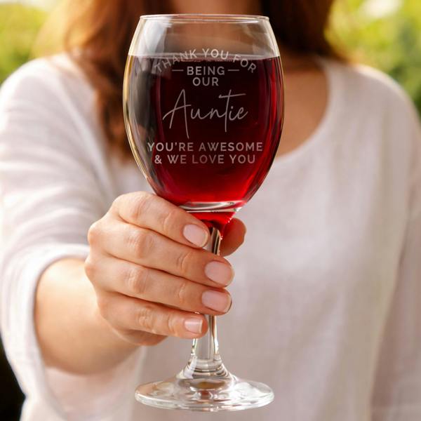 A woman holding up a large wine glass filled with red wine. The glass is laser-engraved with the words "Thank You For Being Our Auntie, You're Awesome & We Love You" in a mix of modern sans-serif and elegant script fonts.