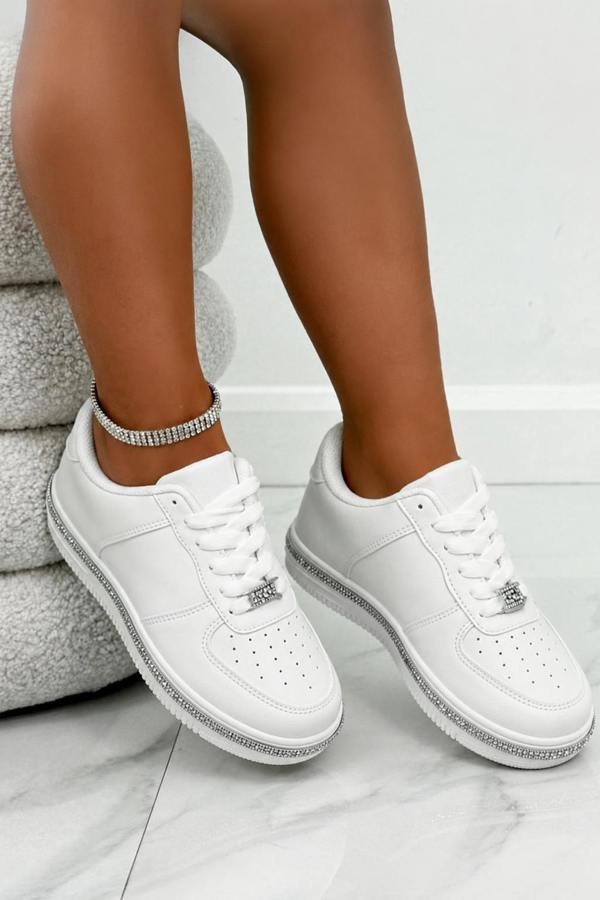 A pair of white low-top sneakers with silver rhinestone trim along the midsoles and silver ankle bracelets worn with them, shown on a model's feet.