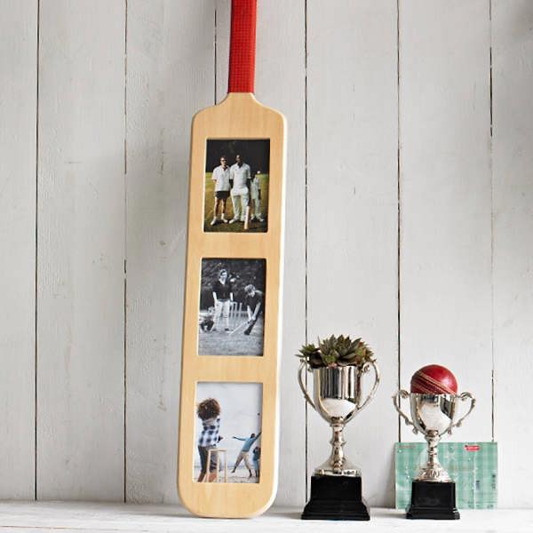 A wooden cricket bat-shaped photo frame featuring three vertical photos, displayed standing upright on a white wooden surface next to two silver trophies and a cricket ball.