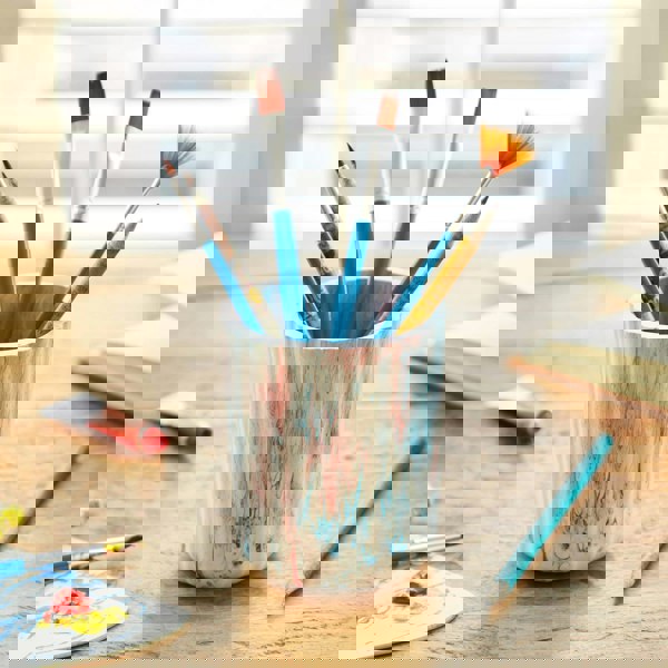 A tumbler of paint brushes on a wooden table.