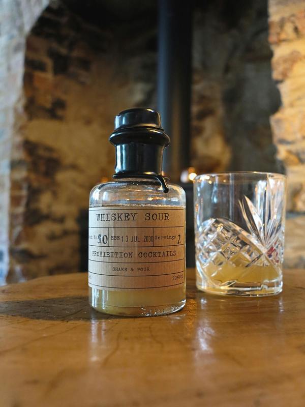 A bottle of Whiskey Sour cocktail mix with a black cap and cream-colored label, placed on a wooden table next to a clear cut-glass rocks tumbler containing a golden liquid, with a rustic stone archway in the background.