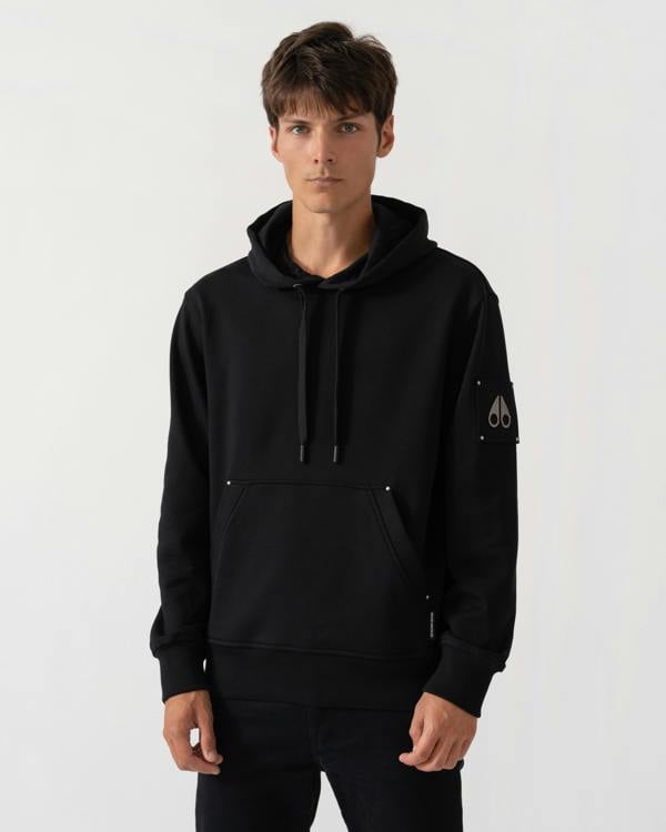 A man wearing a black pullover hoodie with drawstrings, a kangaroo pocket, and a small white embroidered logo on the left sleeve, photographed against a light gray studio background.