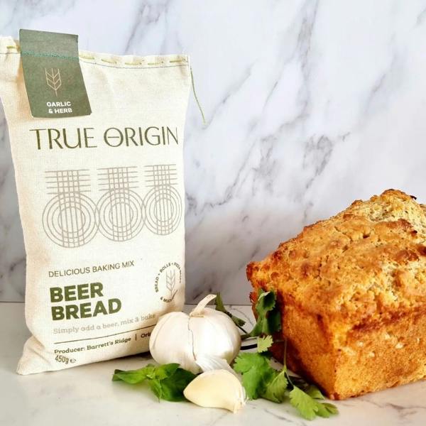 A fabric bag of True Origin Garlic and Herb Beer Bread mix next to a whole garlic bulb, loose cloves, fresh green herbs, and a crusty golden bread loaf against a white marble background.