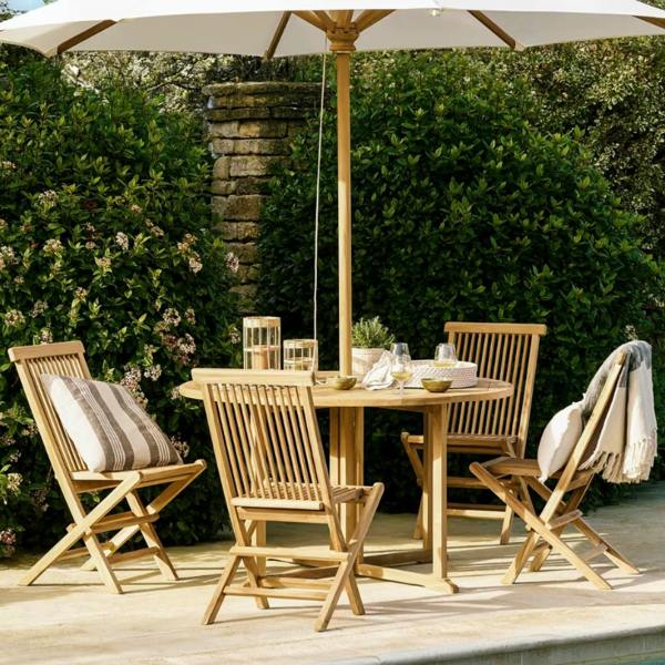 A 3m wooden teak garden parasol with an open natural white canopy shading a round timber dining table and folding chairs next to a swimming pool edge.
