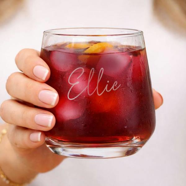 A close-up of a personalised name tumbler glass etched with the name Ellie in a script font, filled with a red iced cocktail and citrus wheels.