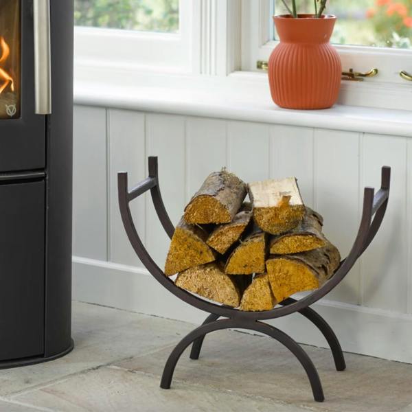 A dark wrought iron curved log holder stacked with firewood logs on a stone floor next to an indoor wood burner.