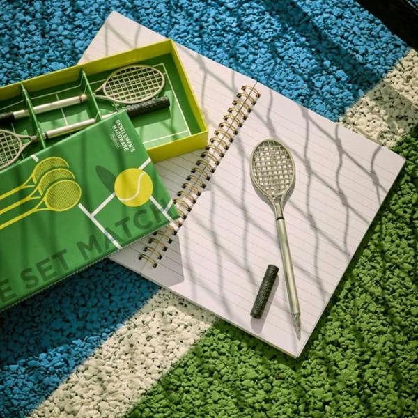 A novelty tennis racket ballpoint pen uncapped next to an open spiral notebook on a blue and green tennis court surface alongside its themed gift box.