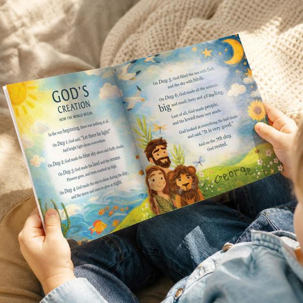 A top-down lifestyle photograph of a young child holding open a copy of the Personalised Children's Bible Stories Book from Abode Home & Gifts, showing a colorful 32-page double spread titled 'God's Creation' with the name George integrated into the grass illustration.