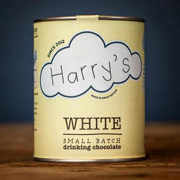 Recyclable soft yellow tin of Harry's Small Batch White Drinking Chocolate resting on a rustic wooden table.