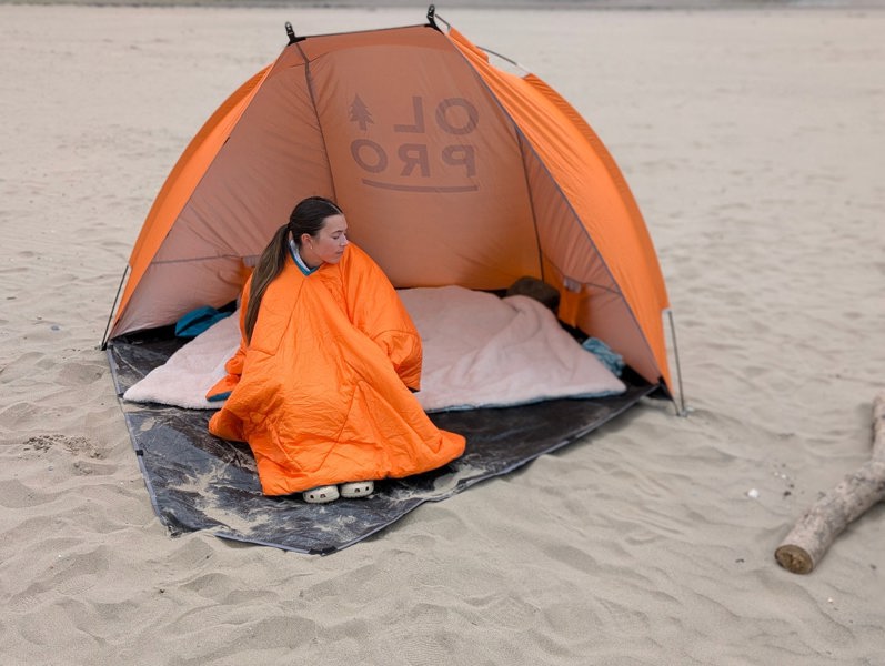 Someone using the OLPRO Insulated Outdoor Reversible Blanko on the beach whilst sat inside of a beach tent.