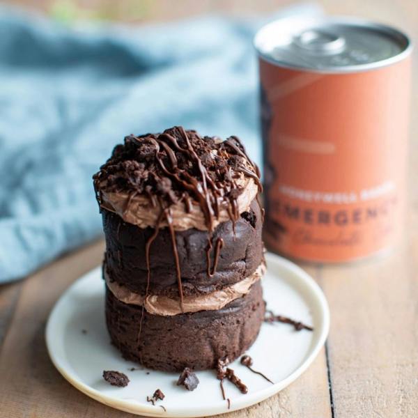 Freshly baked two-tier mini chocolate cake drizzled with dark chocolate frosting, sitting on a small ceramic plate next to an orange Emergency Chocolate Cake Kit tin.