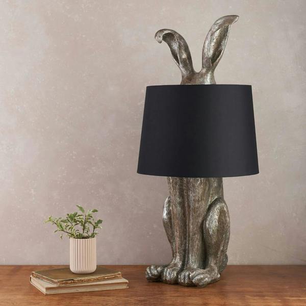 metallic, textured hare-shaped table lamp sitting on a wooden surface, topped with a plain black lampshade. To the left, there is a small potted plant sitting on a stack of old books.