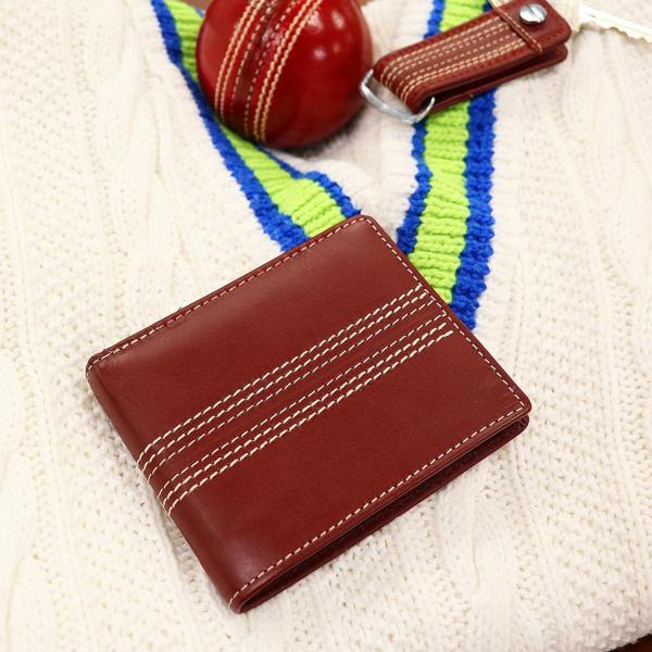 Close-up of a personalised vintage brown leather bi-fold wallet featuring stitched seam lines resembling a cricket ball. The wallet is placed on a white cable-knit cricket sweater next to a red cricket ball and a brown leather keyring. Ideal gift for a cricket fan or player.
