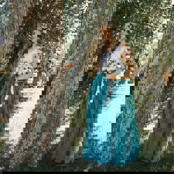 Young girl in a white top and teal skirt standing next to a tree in a park.