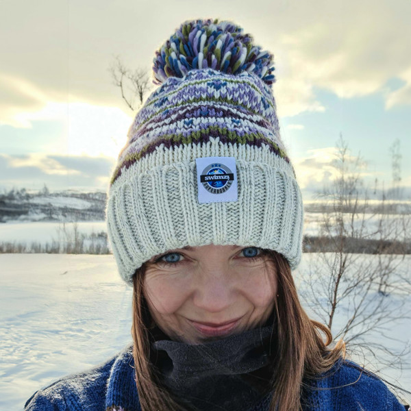 Woman in the Norwegian sun fading over the horizon is wearing a white based bobble hat with heather and moss green traditional fairisle pattern.  The hat also has a deep ribbed headband and contains reflective yarns and a luxury sherpa fleece lining.