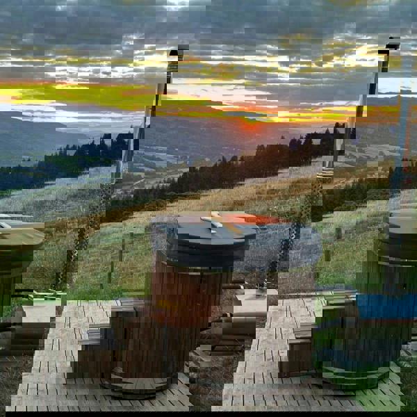 Wood-fired hot tub on a wooden platform with a scenic sunset over mountains and fields.