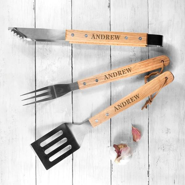 An overhead view of three barbecue utensils including grilling tongs, a fork, and a spatula with timber handles custom-engraved with the name Andrew, arranged on white wooden planks next to a garlic bulb.