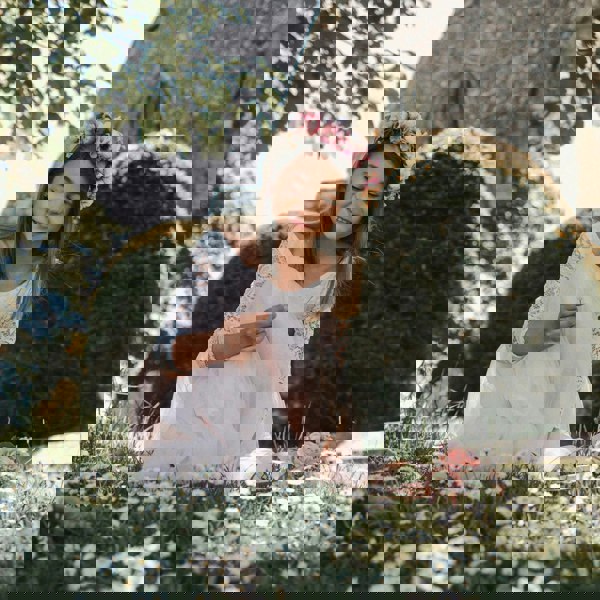 Young girl in a white dress with floral headband sitting on grass with flowers.