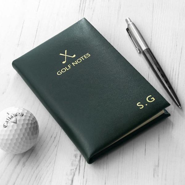 Premium dark green leather golf notebook embossed with the initials 'S.G.' and a gold crossed-golf-club icon, resting on a white surface next to a Callaway golf ball and a silver pen.