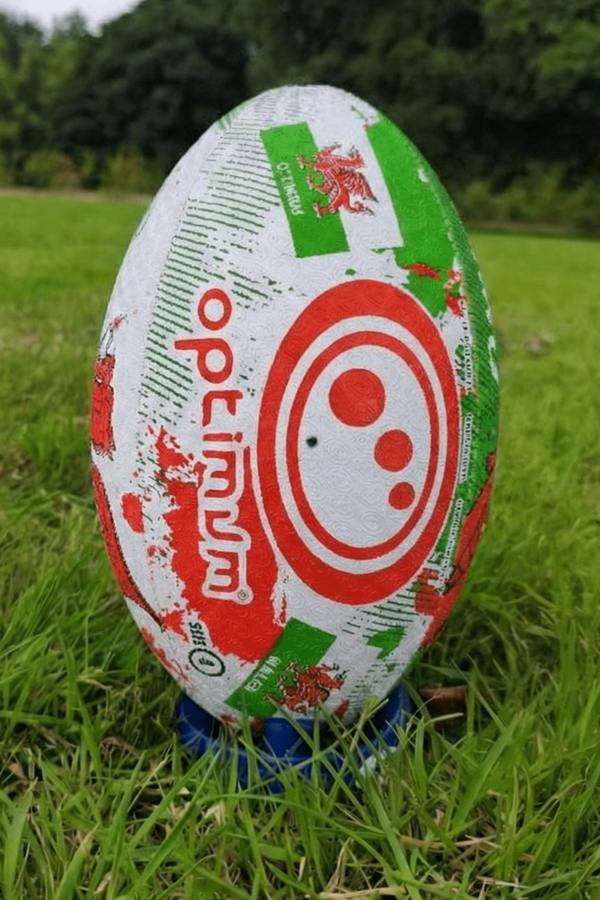 White, red and green Optimum Wales rugby ball showing red Optimum logo, upright on a kicking tee - Optimum