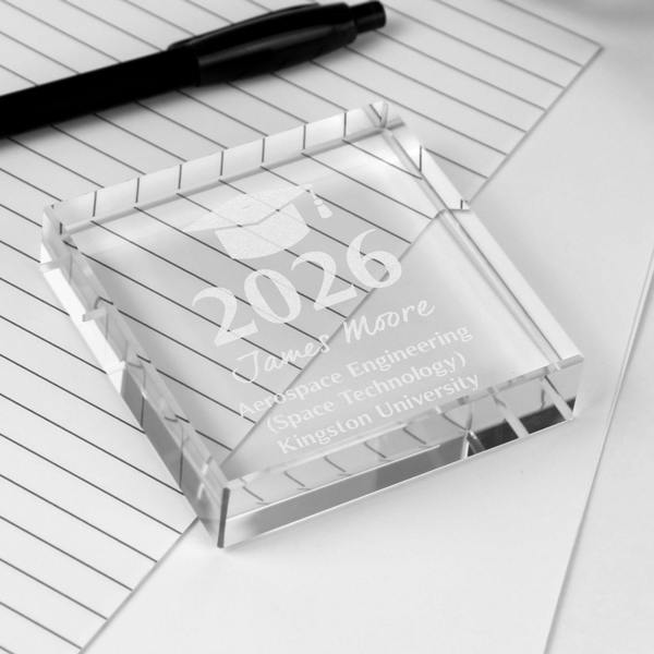 A personalised graduation crystal token engraved with the name James Moore, the year 2026, and an Aerospace Engineering degree from Kingston University, lying flat on lined paper with a pen.