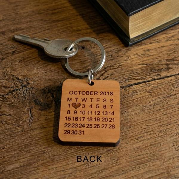 The reverse side of a square cherry wood keyring resting on a rustic old wooden table next to a vintage book and a metal key. The wooden surface is engraved with an October 2018 calendar grid, featuring a small heart symbol marking a specific date, with the word 'BACK' printed below.