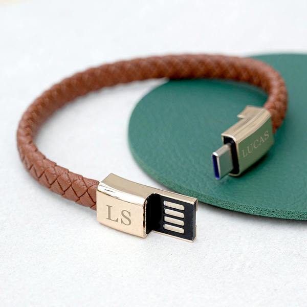 A braided tan leather men's charger bracelet loop with its gold-toned USB-C terminal plug engraved with MH plugged into a laptop port.