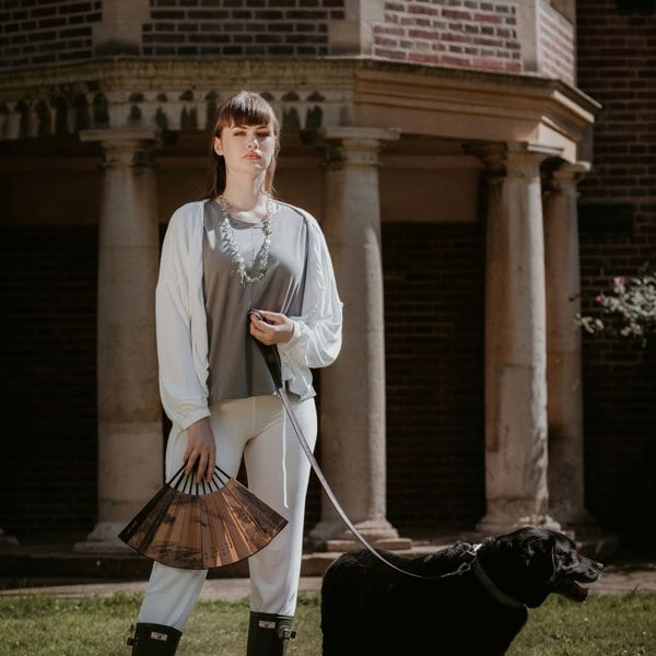 This is a young woman standing in a country garden. She has straight dark hair and is facing the camera. She is holding an open fan in her right hand and she is wearing green wellington boots. She has a black dog on a lead in her left hand. She is wearing a short sleeve tee in steel with a white jacket and trousers.