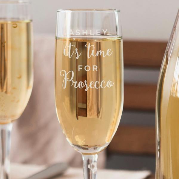 A clear glass champagne flute filled with bubbling prosecco sits on a table. The front of the glass is engraved with the name 'ASHLEY' in capital block letters, above the fixed script phrase 'it's time for Prosecco'. Another blurred flute and a prosecco bottle frame the background.