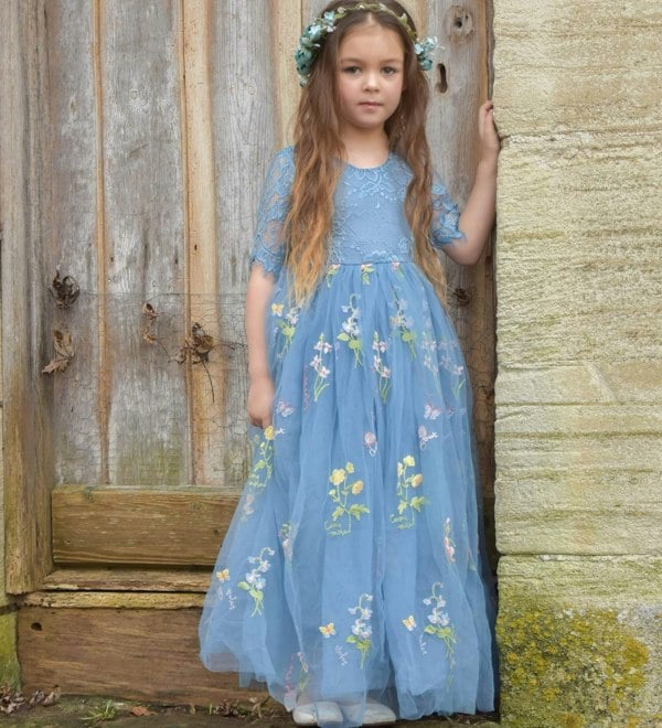 Young girl in a blue floral dress standing against a rustic wooden wall.