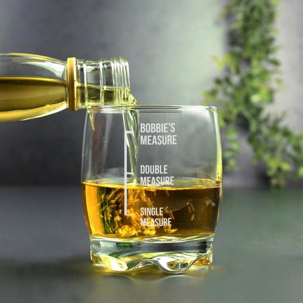 A hand pouring golden whisky from a bottle into a personalised engraved glass tumbler that features measures for "Single Measure", "Double Measure", and "Bobbie's Measure".
