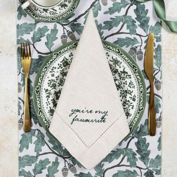 A natural linen-cotton napkin folded neatly on a green botanical leaf plate, showing dark green embroidery that reads 'you're my favourite'.