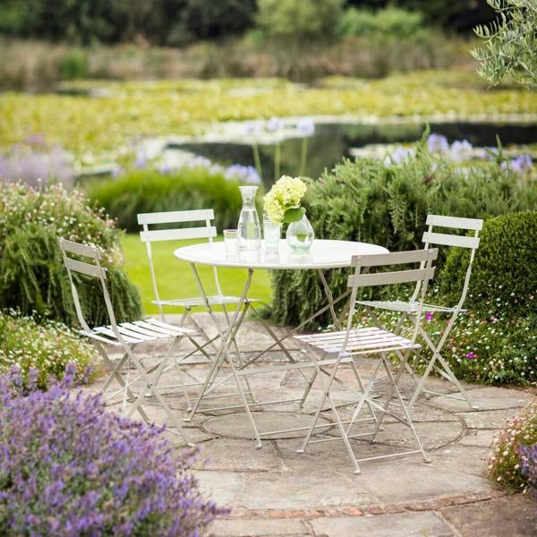 A French-inspired light clay metal round bistro table and four folding chairs set up on a stone garden patio overlooking a pond.