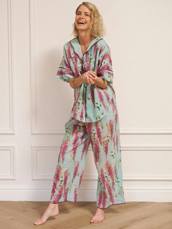 Woman wearing duck egg blue cotton gauze oversized pyjamas with pink wisteria floral print, short-sleeve button-through top and wide-leg trousers, standing barefoot indoors.